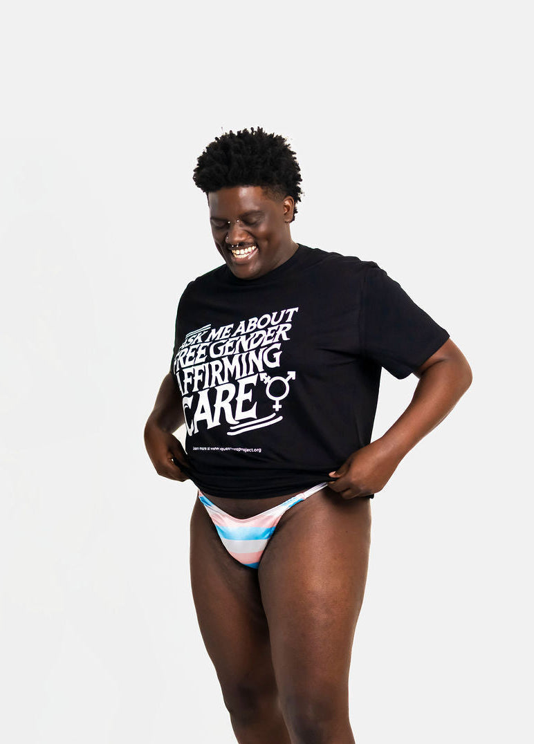 Trans person wearing a black t-shirt that says "ask me about free gender affirming care" while modeling a tucking gaff on a white background