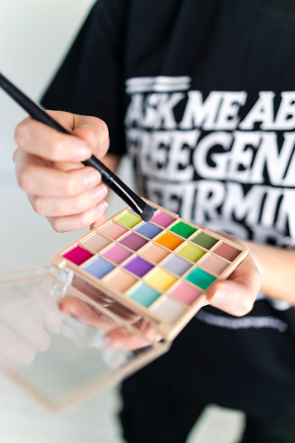 Person holding a colorful eyeshadow palette with a brush, wearing a black shirt with white text.